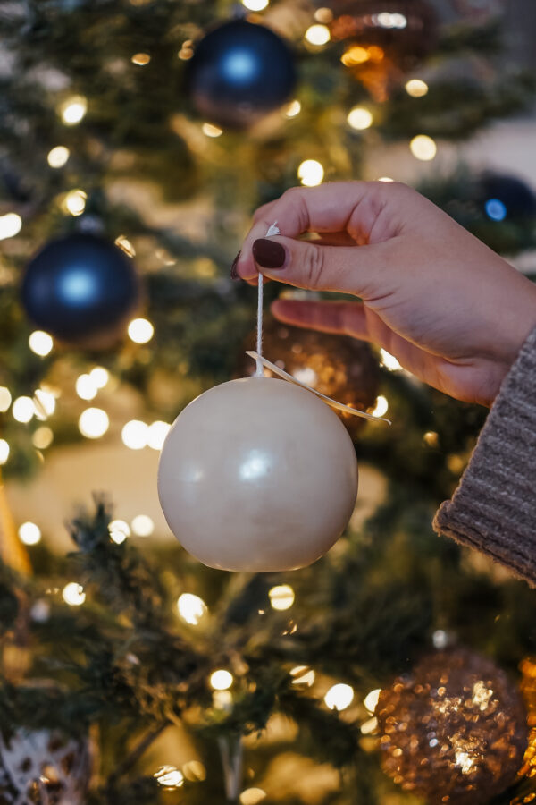 Bougie blanche en forme de boule de Noël tenue à la main devant un sapin décoré